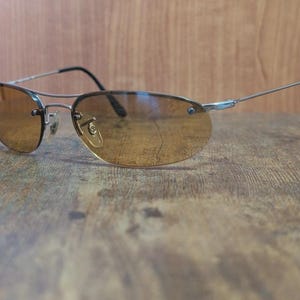 May include: Rimless sunglasses with a silver metal frame and brown tinted lenses. The sunglasses have a classic aviator shape with a double bridge and black earpieces. The lenses are oval-shaped and provide a warm, amber hue.