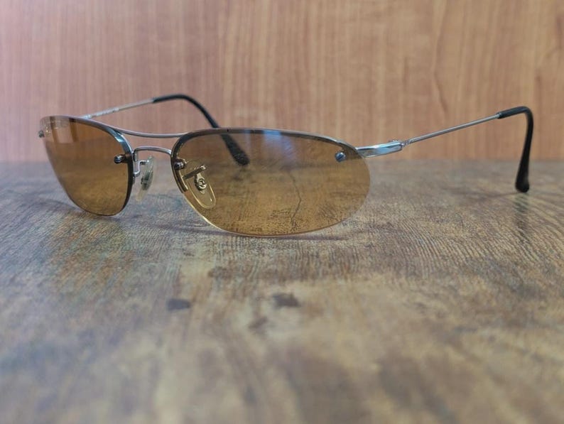 May include: Rimless sunglasses with a silver metal frame and brown tinted lenses. The sunglasses have a classic aviator shape with a double bridge and black earpieces. The lenses are oval-shaped and provide a warm, amber hue.