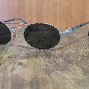 May include: Oval-shaped sunglasses with dark lenses and a silver metal frame. The temples have a patterned design in shades of blue and gray. The sunglasses are resting on a wooden surface.