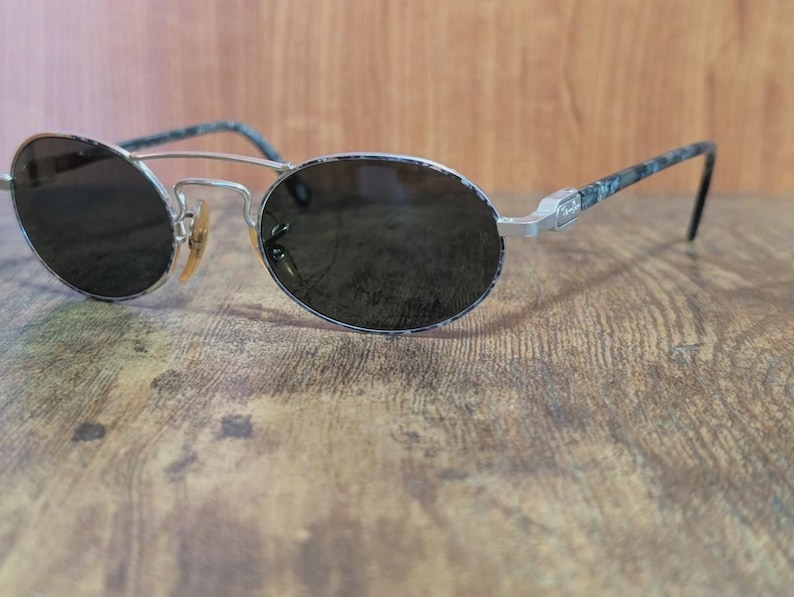 May include: Oval-shaped sunglasses with dark lenses and a silver metal frame. The temples have a patterned design in shades of blue and gray. The sunglasses are resting on a wooden surface.