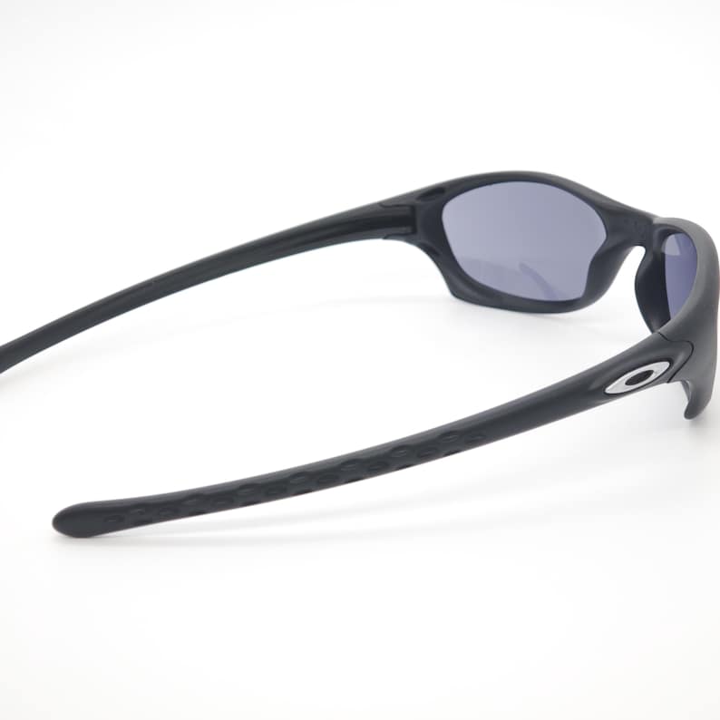 May include: Black sunglasses with gray lenses. The sunglasses have a sleek, curved design with textured arms and a small silver logo on the side. The lenses are a dark gray color, and the frame is matte black.