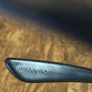 May include: Close-up of a black eyeglass arm with the engraved text "W1955 WYAO". The arm is angled, with a smooth, matte finish. The background is a wooden surface.