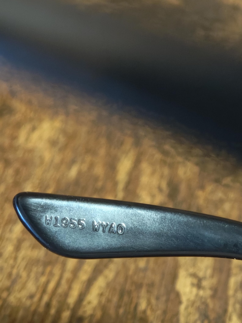 May include: Close-up of a black eyeglass arm with the engraved text "W1955 WYAO". The arm is angled, with a smooth, matte finish. The background is a wooden surface.