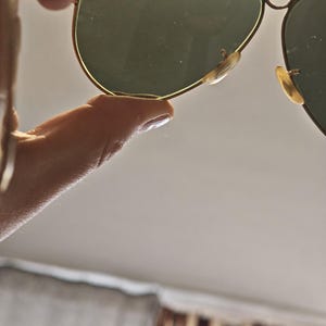 May include: Gold-framed aviator sunglasses with green-tinted lenses. The sunglasses have a classic teardrop shape and are held up by a hand. The nose pads are a light beige color.