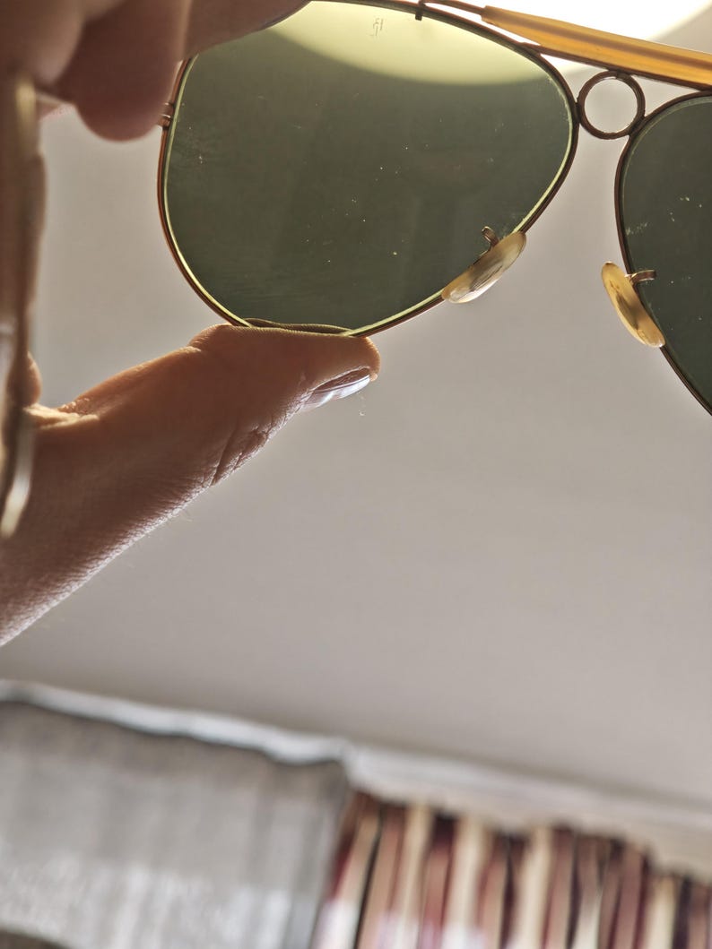 May include: Gold-framed aviator sunglasses with green-tinted lenses. The sunglasses have a classic teardrop shape and are held up by a hand. The nose pads are a light beige color.