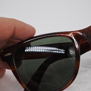 May include: Tortoiseshell sunglasses with green lenses. The frames have a classic design with a subtle pattern. The sunglasses are held by a hand, suggesting they are being showcased or examined.