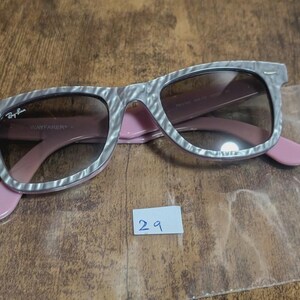 Puede incluir: Gafas de sol Ray-Ban Wayfarer con montura estampada gris y blanca, lentes marrones y patillas rosas. Las gafas de sol est&aacute;n sobre una superficie de madera. El logotipo de Ray-Ban es visible.