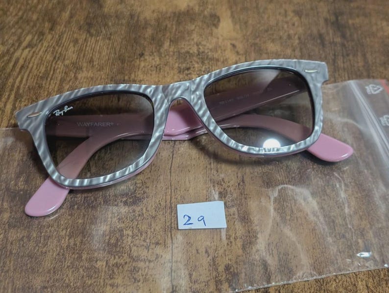 Puede incluir: Gafas de sol Ray-Ban Wayfarer con montura estampada gris y blanca, lentes marrones y patillas rosas. Las gafas de sol est&aacute;n sobre una superficie de madera. El logotipo de Ray-Ban es visible.