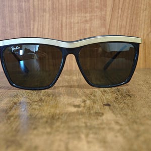 Peut inclure: Lunettes de soleil noires avec une monture sup&eacute;rieure dor&eacute;e et des verres fonc&eacute;s. Les lunettes de soleil ont une forme carr&eacute;e et un logo Ray-Ban sur la lentille gauche. Elles sont pos&eacute;es sur une surface en bois.