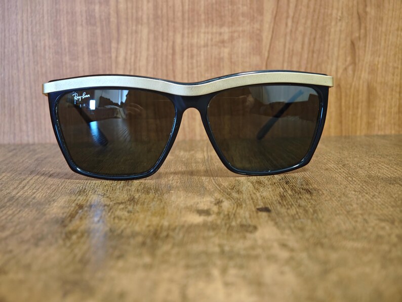 Peut inclure: Lunettes de soleil noires avec une monture sup&eacute;rieure dor&eacute;e et des verres fonc&eacute;s. Les lunettes de soleil ont une forme carr&eacute;e et un logo Ray-Ban sur la lentille gauche. Elles sont pos&eacute;es sur une surface en bois.