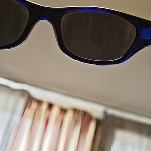 May include: Close-up of black sunglasses with dark lenses and blue accents on the temples. The sunglasses have a rectangular shape and a white trim along the top edge of the frames. The lenses appear to be tinted.