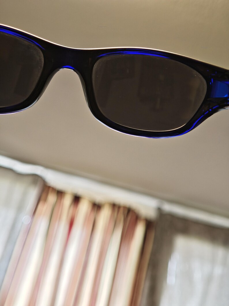 May include: Close-up of black sunglasses with dark lenses and blue accents on the temples. The sunglasses have a rectangular shape and a white trim along the top edge of the frames. The lenses appear to be tinted.