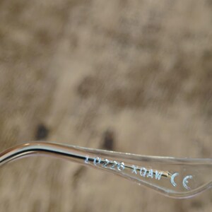 Puede incluir: Primer plano de la patilla de unas gafas. La patilla es de pl&aacute;stico transparente con detalles met&aacute;licos plateados. El texto "L0226 XQAW CE" est&aacute; impreso en la patilla en color azul claro. El fondo es borroso y de tono neutro.