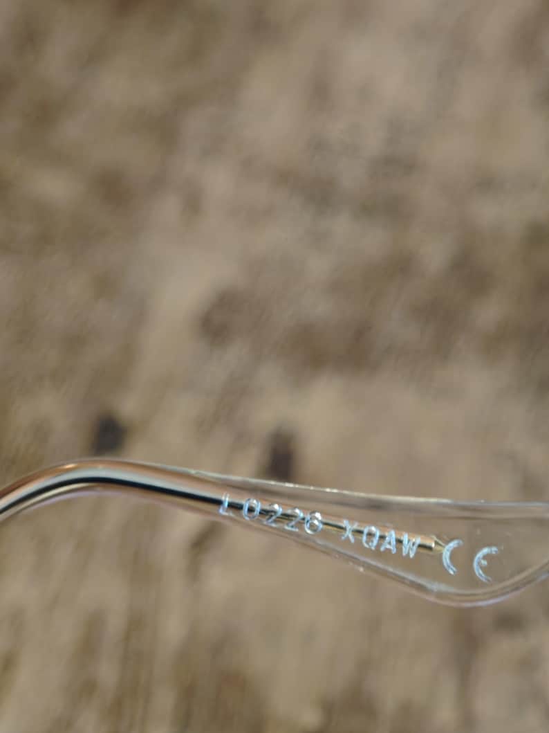 Puede incluir: Primer plano de la patilla de unas gafas. La patilla es de pl&aacute;stico transparente con detalles met&aacute;licos plateados. El texto "L0226 XQAW CE" est&aacute; impreso en la patilla en color azul claro. El fondo es borroso y de tono neutro.