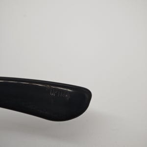 May include: Close-up of a black eyeglass frame arm with the word "OPTICS" printed on it. The frame is made of a glossy material and is set against a plain white background.