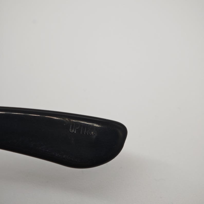 May include: Close-up of a black eyeglass frame arm with the word "OPTICS" printed on it. The frame is made of a glossy material and is set against a plain white background.