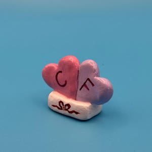 Könnte beinhalten: Kleine, handbemalte Skulptur mit zwei verbundenen Herzen. Ein Herz ist rosa mit einem schwarzen „C“, das andere ist lila mit einem schwarzen „F“. Die Herzen ruhen auf einem weißen Sockel mit einem dekorativen Wirbel. Der Hintergrund ist einfarbig blau.