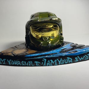May include: A green and gold helmet figurine, inspired by a science fiction character, rests on a decorative base. The base is painted with blue and black details and features the text "I AM YOUR SHIELD I AM YOUR SWORD."