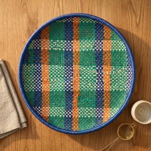 May include: A round woven basket with a blue rim and a colorful plaid pattern. The basket features vertical stripes of orange and horizontal stripes of green and blue. The basket is on a wooden surface.