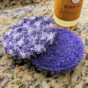 May include: Two handmade, round dish scrubbers. One is solid purple, the other is a mix of purple and white. The scrubbers are made of a textured, looped yarn. A bottle of dish soap is in the background.