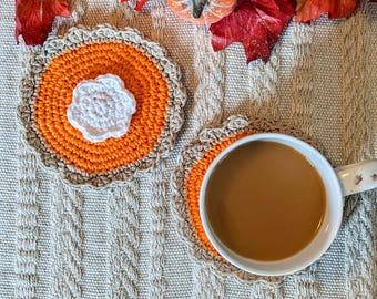 May include: Two handmade crochet coasters in the shape of pumpkin pies. The coasters feature orange centers, tan crusts, and white floral accents. One coaster holds a white mug filled with a brown beverage. The coasters are on a textured beige surface.