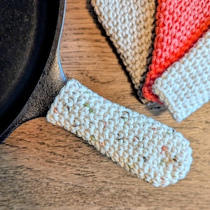 May include: A cast iron skillet with a cream-colored, speckled, knitted handle cover. Several other knitted handle covers in white, coral, and tan are visible. The items are on a wooden surface. Kitchen accessories.