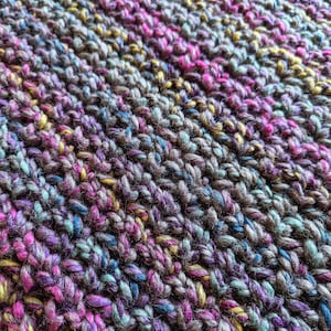 May include: Close-up of a knitted fabric with a multi-colored striped pattern. The fabric is made with a variety of shades of pink, purple, blue, green, and brown yarn.