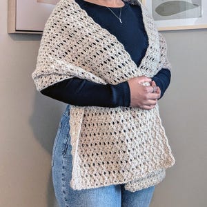 May include: A beige crochet shawl with a starburst pattern. The shawl is draped over a person wearing a black long-sleeved shirt and blue jeans. The shawl has a rectangular shape with a fringed edge.
