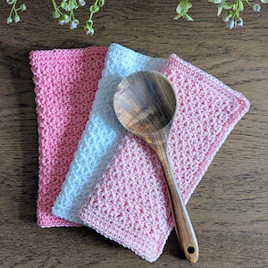 Puede incluir: Una cuchara de madera descansa sobre tres paños de cocina de ganchillo en rosa y blanco. Los paños están colocados sobre una superficie de madera, con una ramita de vegetación en el fondo. La cuchara es de madera y tiene un agujero en el mango.