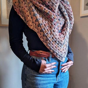 May include: A multi-colored crocheted shawl in shades of brown, pink, and blue. The shawl is draped over a black long-sleeved shirt and blue jeans. A brown leather belt is worn at the waist.