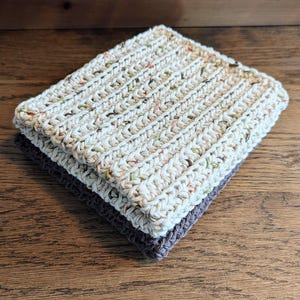 May include: Two handmade crochet washcloths. The top washcloth is off-white with flecks of colour, and the bottom washcloth is a solid dark grey. The washcloths are stacked, showing the texture of the crochet.