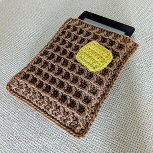 May include: A brown crocheted e-reader case with a yellow crocheted square in the center.