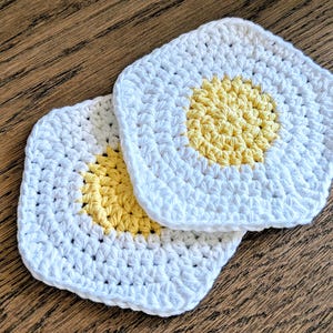 May include: Two handmade, square crochet coasters. Each coaster features a white border and a yellow center, resembling a fried egg. The coasters are placed on a wooden surface, showcasing the texture and color of the yarn.