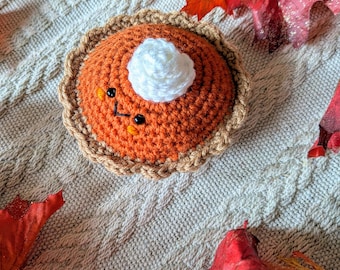 May include: A crocheted pumpkin pie with a smiling face and whipped cream topping. The pie is orange with a tan crust and a white dollop of cream. The pie is on a cream-colored knitted surface with fall leaves.