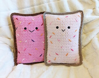 May include: Two crocheted pillows shaped like a slice of toast with pink and white frosting and sprinkles. The pillows have embroidered eyes and a smiling mouth.