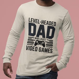 Puede incluir: Una camiseta de manga larga de color beige claro con el texto "LEVEL-HEADED DAD WHO LOVES VIDEO GAMES" en negro. El diseño incluye un gráfico de mando. La camiseta la lleva una persona.