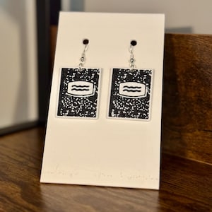 Puede incluir: Un par de pendientes rectangulares colgantes con un diseño de portada de cuaderno de notas con motas blancas y negras. Cada pendiente tiene un borde blanco y una etiqueta central blanca con tres líneas onduladas horizontales.