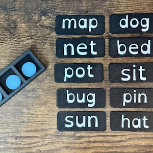 May include: A set of black rectangular tiles with white text reading map, dog, net, bed, pot, sit, bug, pin, sun, and hat. Beside the tiles is a black rectangular frame with three circular light blue tokens inside for phonics learning.