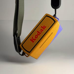 May include: A gold and purple Kodak camera case with a black border and the Kodak logo in red. The case is attached to a green strap and black clasp.