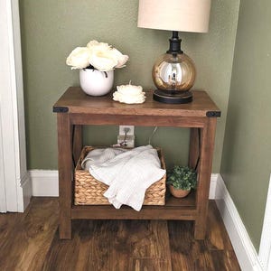 May include: A wooden side table with a lower shelf and a woven basket containing a white blanket. The tabletop holds a white vase with white flowers, a candle, and a lamp with a beige shade and a round, amber-colored base.