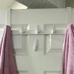 May include: A white over-the-door towel rack with four hooks, holding two pink towels. The rack is mounted on a white door. A white adhesive hook is also attached to the door.