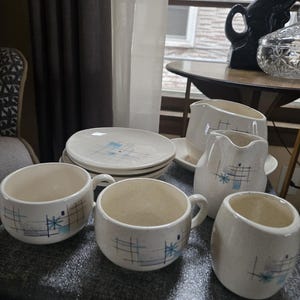 May include: A collection of vintage cream-colored dishware with a mid-century modern design. The set includes plates, cups, a creamer, and a sugar bowl. Each piece features a blue geometric pattern.