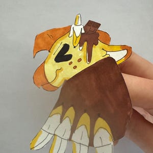 May include: A whimsical paper cutout of a banana split. The banana is yellow with a white peel, topped with a chocolate bar and chocolate sauce. The bottom portion resembles a hand with banana slices.