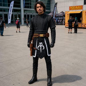 May include: A person in a black costume with white accents, a brown belt, and a holster. The costume features a Mandalorian skull emblem on the front. The person is wearing black gloves and boots. The background includes a food stand and banners.