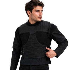 May include: Black tactical vest with quilted shoulder padding and adjustable buckles. The vest is worn over a black long-sleeve shirt. The vest has a utilitarian design.