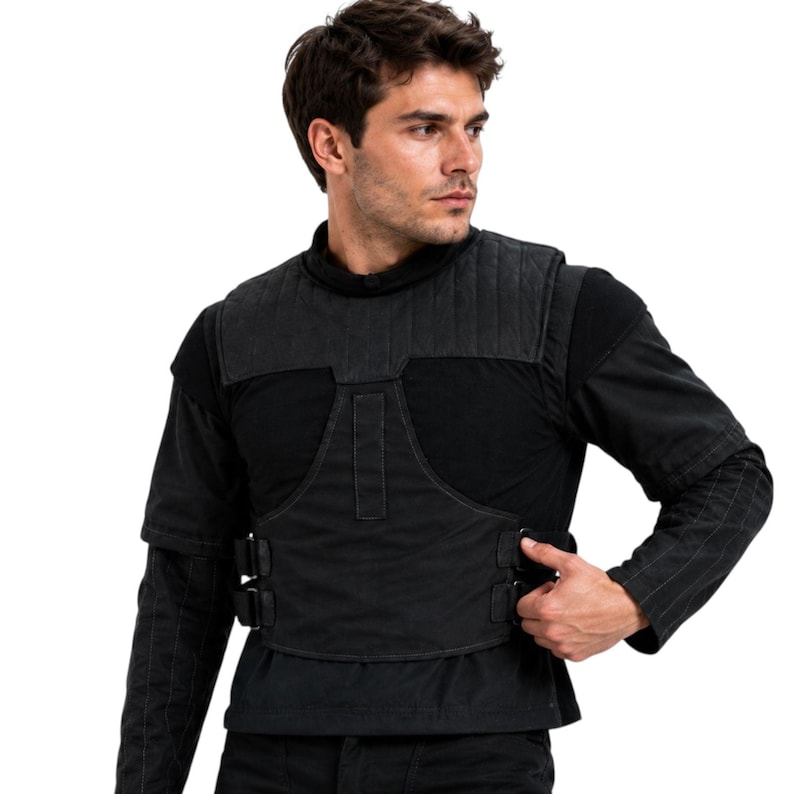 May include: Black tactical vest with quilted shoulder padding and adjustable buckles. The vest is worn over a black long-sleeve shirt. The vest has a utilitarian design.