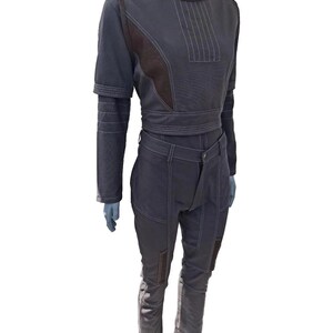 K&ouml;nnte beinhalten: Ein dunkelgraues zweiteiliges Outfit mit wei&szlig;en Nahtdetails. Das Oberteil ist ein kurzes, lang&auml;rmliges Design mit hohem Kragen und braunen Akzenten. Die Hose ist hoch tailliert mit braunen Patches an den Knien.