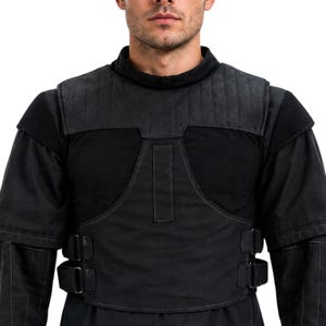 May include: Black padded vest with a high collar and shoulder protection. The vest has a layered design with stitched detailing and adjustable straps. The garment is made of a dark, possibly canvas-like material, and is designed for protection.