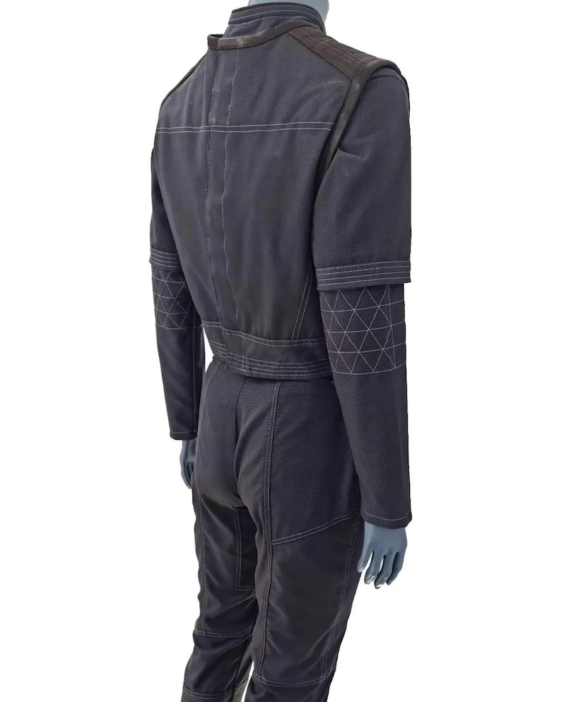 K&ouml;nnte beinhalten: Dunkelgrauer Overall mit wei&szlig;en Steppn&auml;hten. Der Overall hat einen G&uuml;rtel in der Taille, lange &Auml;rmel und geometrische Muster an den Oberarmen. Das Kleidungsst&uuml;ck wird auf einer Schaufensterpuppe pr&auml;sentiert.