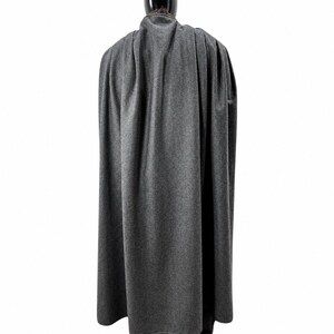 May include: A gray cape made of a soft, textured fabric. The cape is draped over a black mannequin, showcasing its full length and flowing design. The cape's color is a medium gray, and it appears to be made of a wool or similar material.